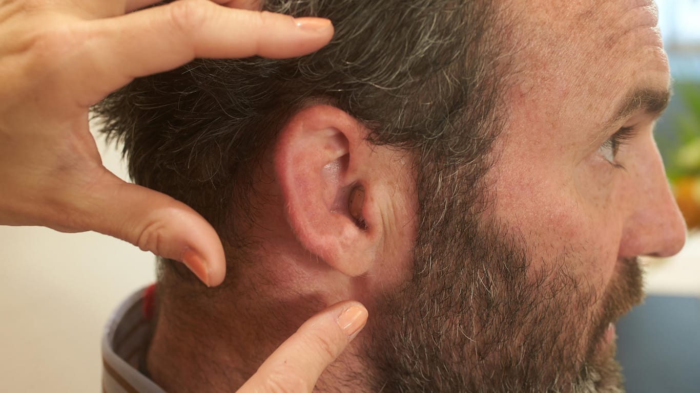 Ein Hand deutet auf das Ohr eines Mannes, in das ein Hörgerät eingesetzt worden ist.
