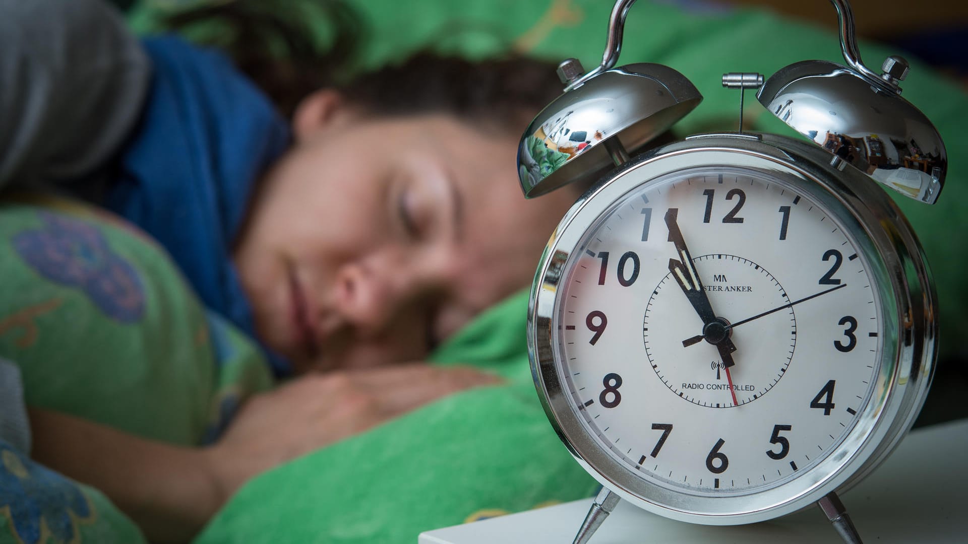 Eine Frau schläft im Bett, während ein Wecker neben ihr auf dem Nachttischränkchen steht.