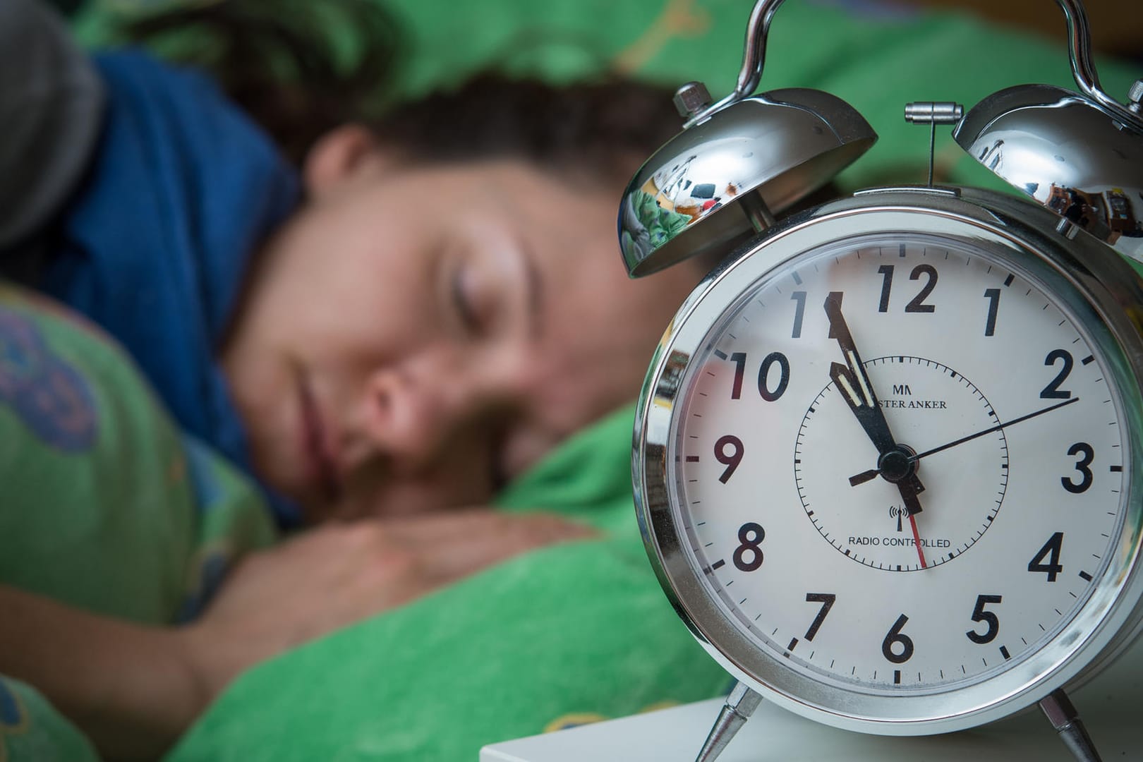 Eine Frau schläft im Bett, während ein Wecker neben ihr auf dem Nachttischränkchen steht.