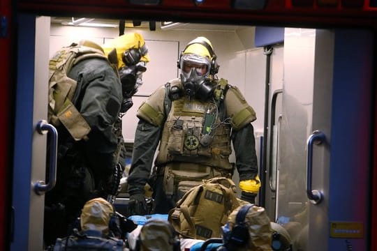 SEK-Beamten mit Atemschutzmasken und Schutzanzügen stehen in einem Fahrzeug vor einem Wohnkomplex.