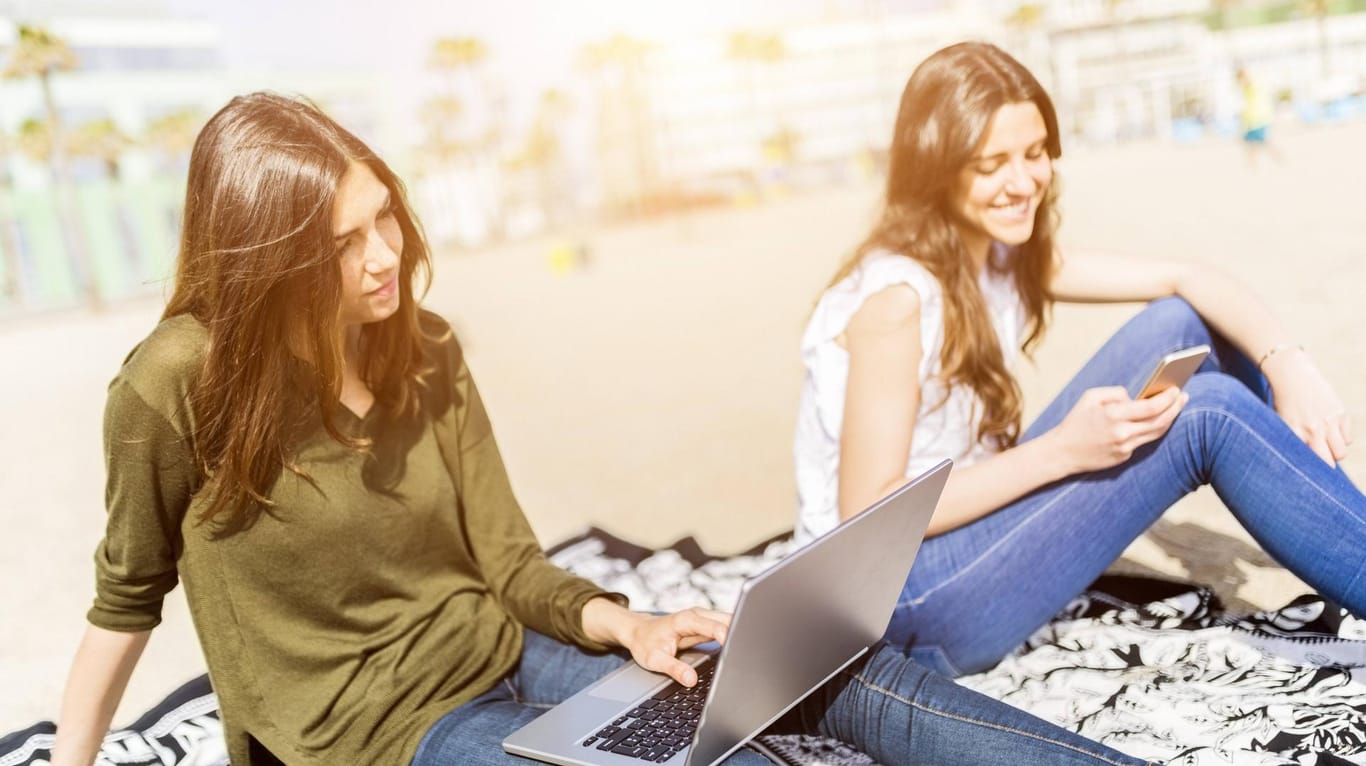 Zwei junge Frauen sitzen mit Laptop und Smartphone am Strand: Nur wenige Urlauber verzichten in den Ferien auf ihre Elektronik.