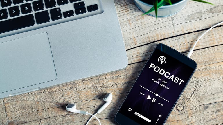 Ein Smartphone mit geöffneter Podcast-App liegen neben einem Laptop auf einem Holztisch.