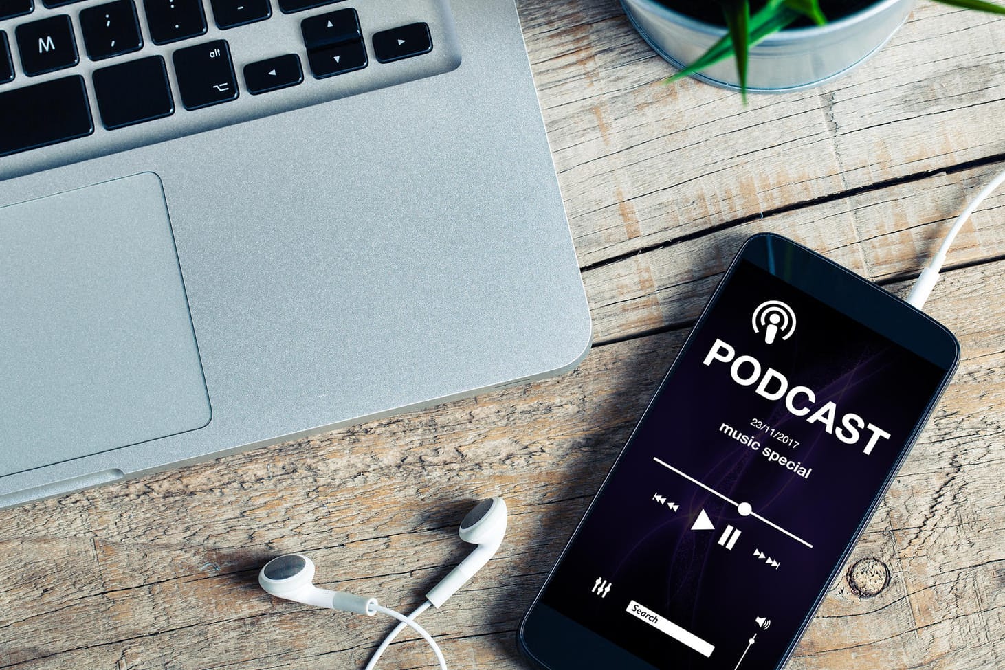 Ein Smartphone mit geöffneter Podcast-App liegen neben einem Laptop auf einem Holztisch.