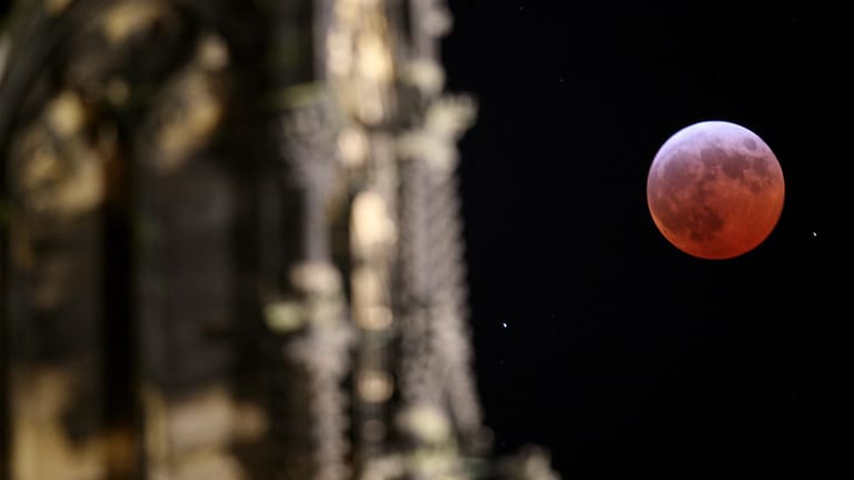 Neben der Fassade des Kölner Domes leuchtet der Mond während einer Mondfinsternis rot bis orange: Die totale Mondfinsternis am späten Freitagabend wird in Nordrhein-Westfalen wahrscheinlich gut zu sehen sein.