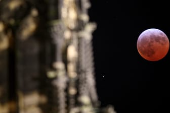 Neben der Fassade des Kölner Domes leuchtet der Mond während einer Mondfinsternis rot bis orange: Die totale Mondfinsternis am späten Freitagabend wird in Nordrhein-Westfalen wahrscheinlich gut zu sehen sein.