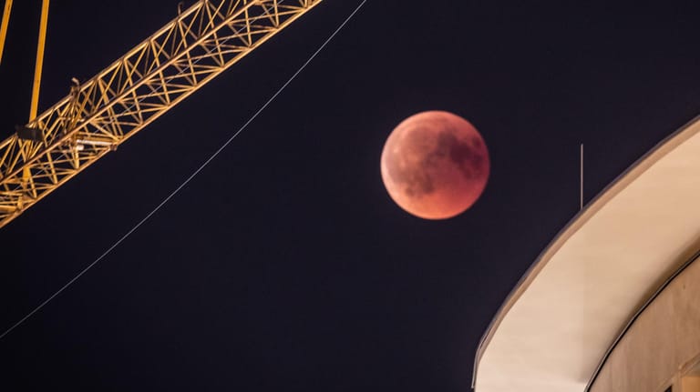 Frankfurt am Main: Zwischen einem Baukran und einem Gebäude in der Innenstadt ist der rötlich gefärbte Vollmond zu sehen: Bei der längsten Mondfinsternis des 21. Jahrhunderts taucht der Mond in den Erdschatten ein.