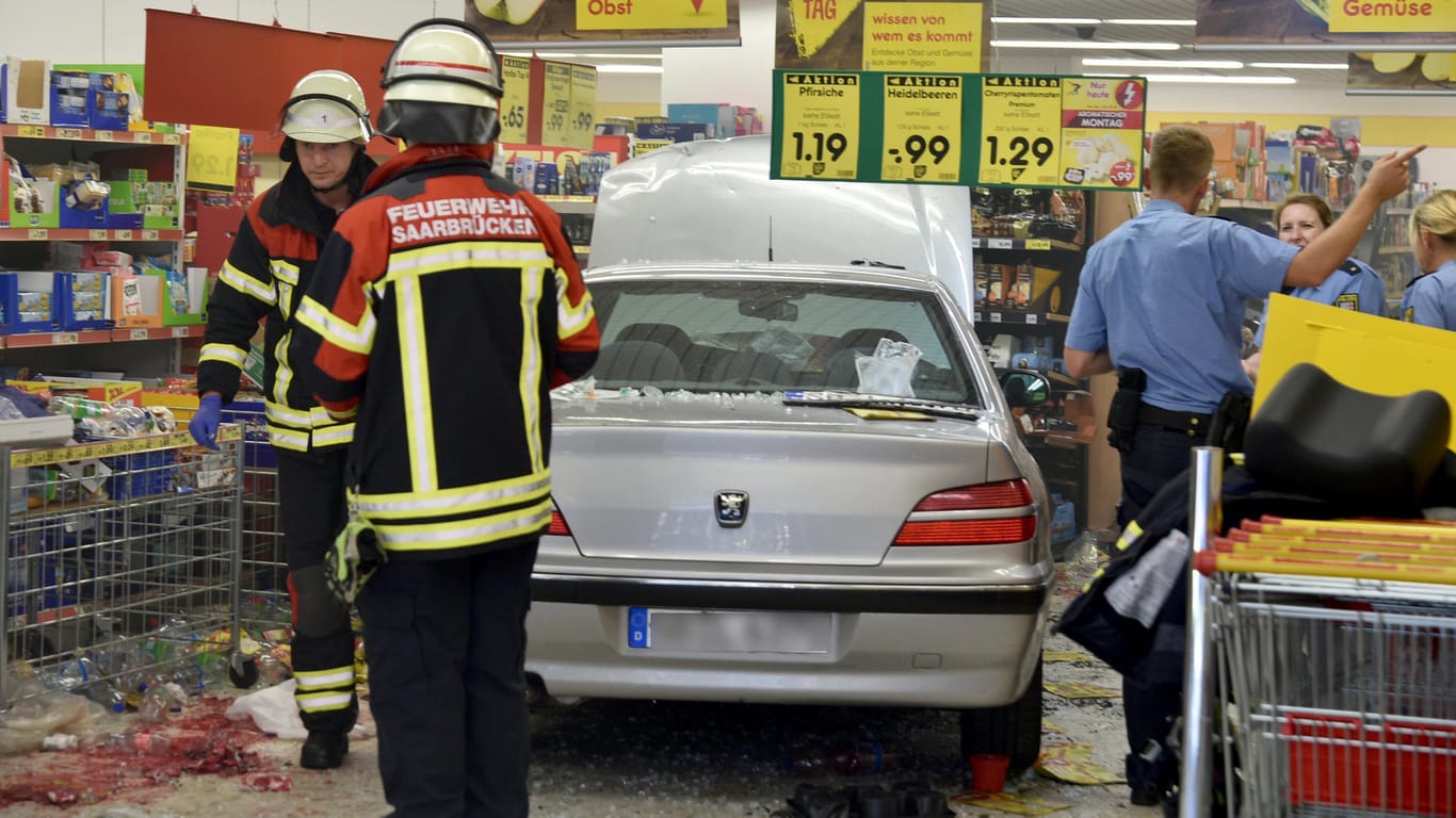 Feuerwehrleute und Polizeibeamte stehen in einem Supermarkt neben einem Pkw: Er war mitten in den Supermarkt gefahren, die Ursache ist noch unklar.