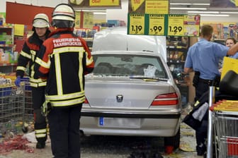 Feuerwehrleute und Polizeibeamte stehen in einem Supermarkt neben einem Pkw: Er war mitten in den Supermarkt gefahren, die Ursache ist noch unklar.