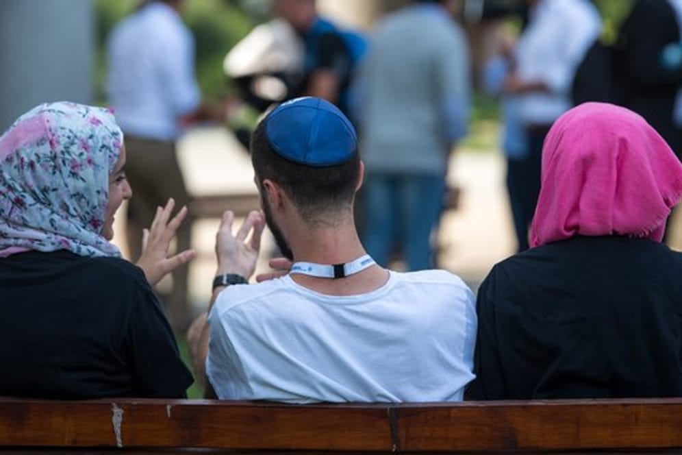 Zwei junge Muslimas mit Kopftuch und ein junger jüdischer Mann mit Kippa unterhalten sich miteinander.