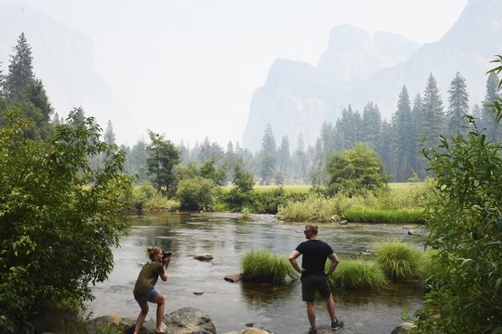 Zwei Touristen am Merced River im Yosemite-Tal, während Rauch im Hintergrund die Bergkulisse verschleiert.