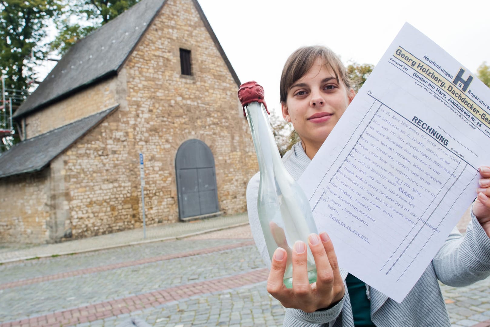 Vanessa Nöhr von der Stadt Goslar zeigt eine alte Flaschenpost sowie eine Kopie des darin gefundenen Schreibens vor der Domvorhalle: Dachdecker hatten die Flasche im Jahr 1930 mit einer Botschaft für die Nachwelt versehen, sie dann aber nicht irgendwo ins Wasser geworfen, sondern unter dem Dach der Domvorhalle versteckt. Dort wurde sie jetzt gefunden.
