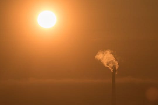 Durch einen Dunstschleier scheint die aufgehende Sonne einen Schornstein an, der seinen Dampf in die Atmosphäre entlässt.
