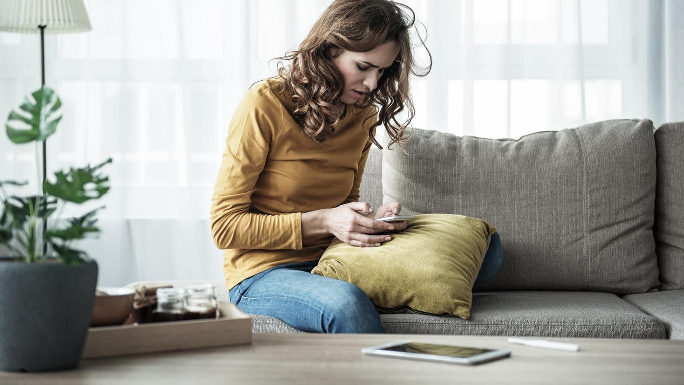 Eine Frau auf dem Sofa blickt besorgt auf ihr Smartphone: Wenn die WLAN-Verbindung im Heimnetzwerk häufig zusammenbricht, liegt der Grund vielleicht im Mauerwerk versteckt.