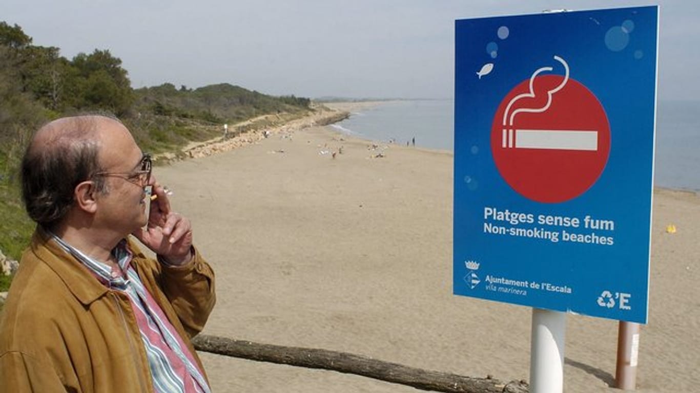 Ein Mann raucht, während er die Aufschrift auf einem Schild am Strand von l'Escala bei Girona im Nordosten Spaniens liest.