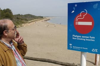 Ein Mann raucht, während er die Aufschrift auf einem Schild am Strand von l'Escala bei Girona im Nordosten Spaniens liest.
