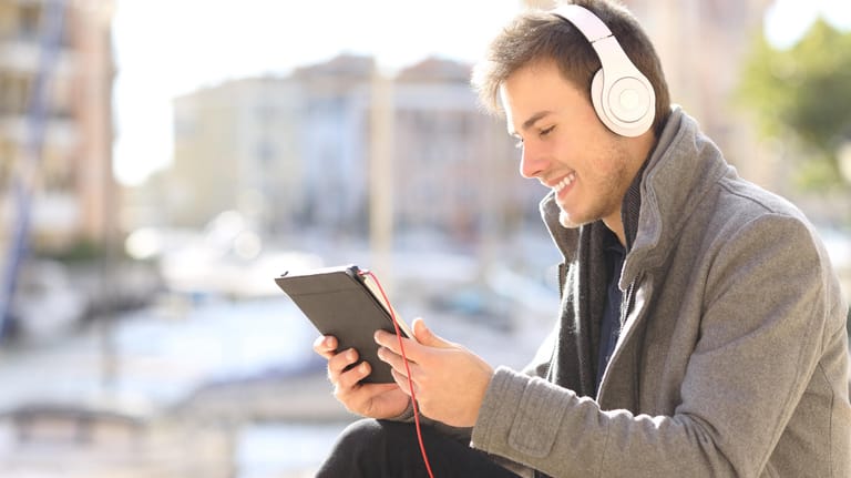 Ein junger Mann sitzt mit Kopfhörern draußen und schaut auf sein Tablet.