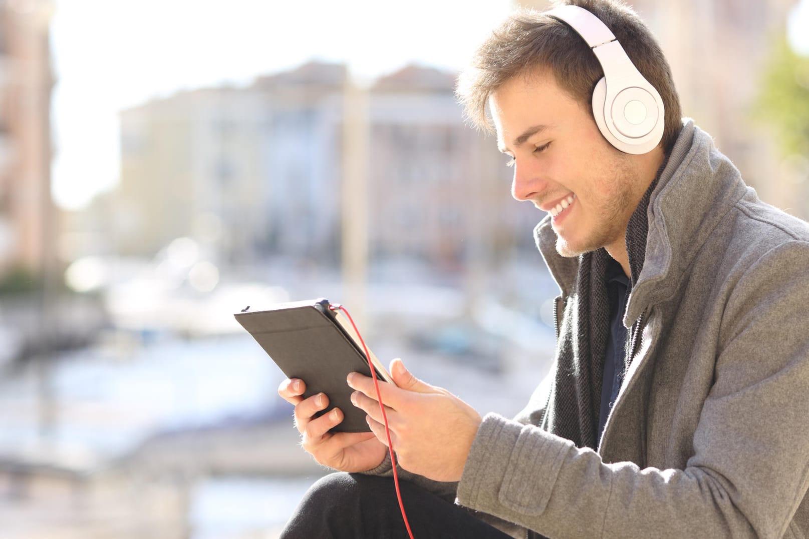 Ein junger Mann sitzt mit Kopfhörern draußen und schaut auf sein Tablet.