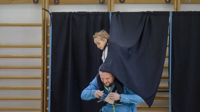 Ein Mann verlässt mit einem Kind auf seinen Schultern eine Wahlkabine. Ein Mann verlässt mit einem Kind auf seinen Schultern eine Wahlkabine.