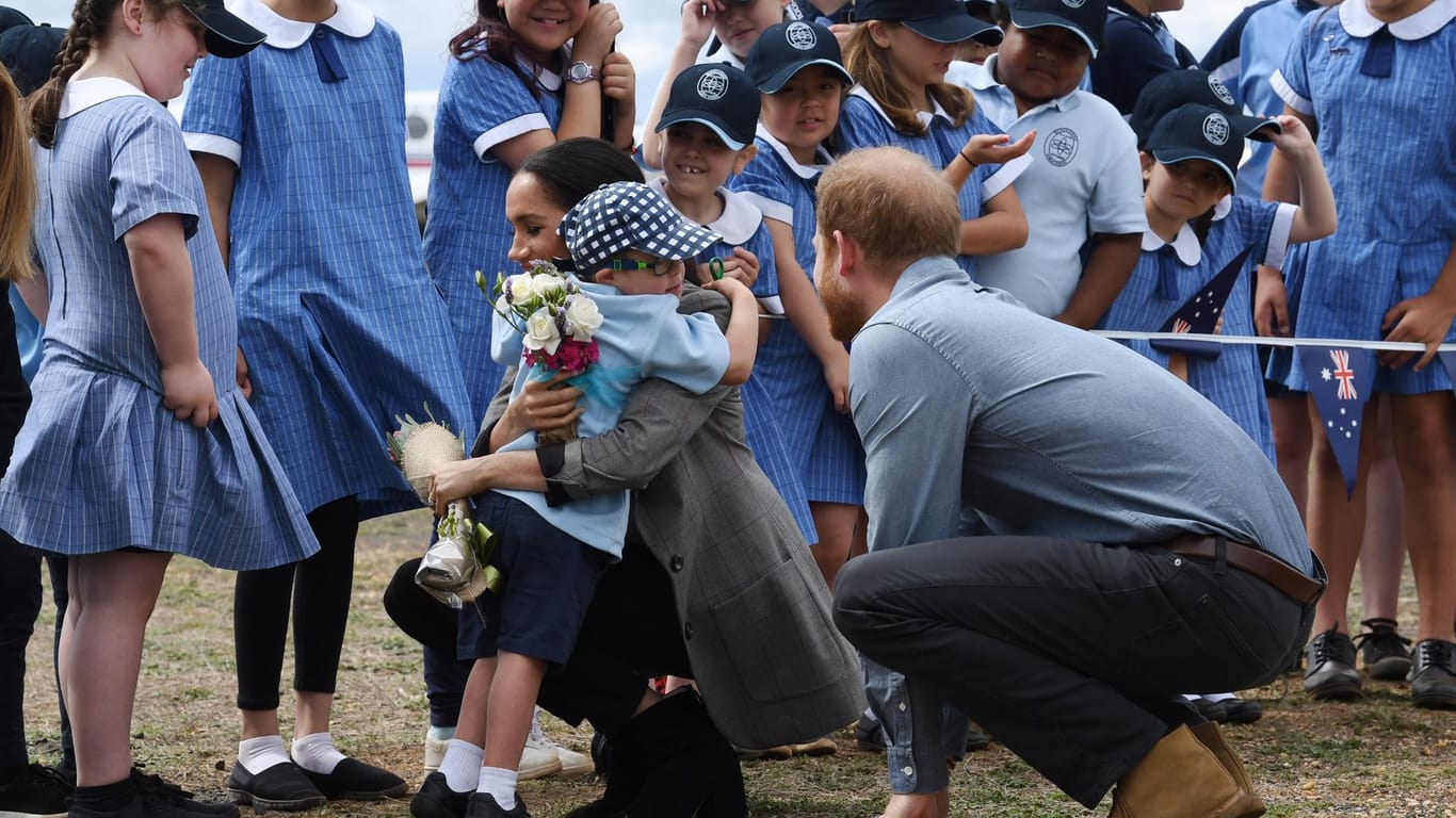 Prinz Harry und Meghan in Australien: Herzogin Meghan (M, r) wird nach der Ankunft am Dubbo Regional Airport vom Schüler Luke Vincent (M, l) von der Buninyong Public School umarmt, während Prinz Harry (r) daneben kniet.