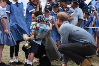 Prinz Harry und Meghan in Australien: Herzogin Meghan (M, r) wird nach der Ankunft am Dubbo Regional Airport vom Schüler Luke Vincent (M, l) von der Buninyong Public School umarmt, während Prinz Harry (r) daneben kniet.