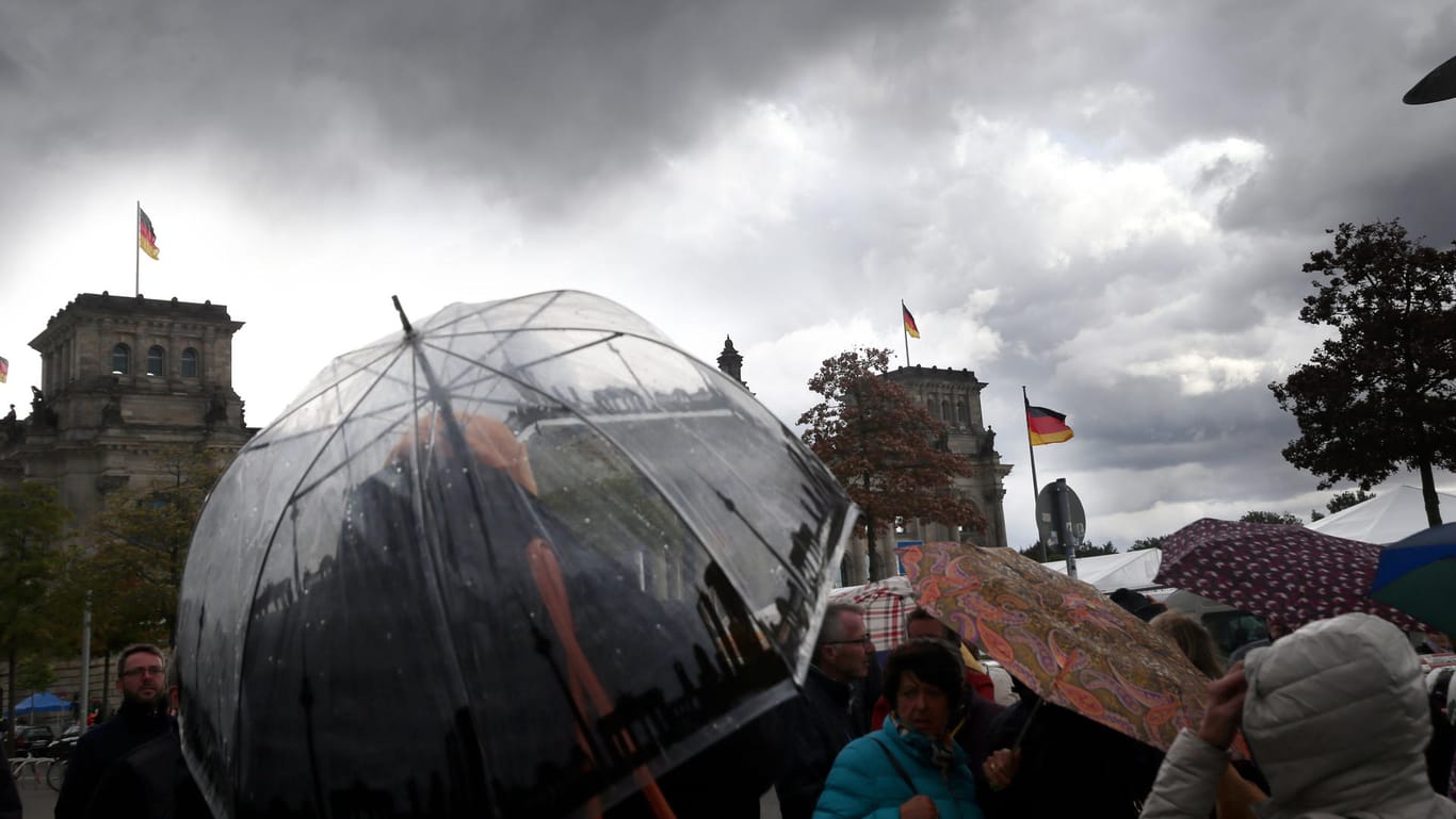 Menschen mit Regenschirmen stehen vor dem Reichstagsgebäude, während dunkle Regenwolken aufziehen: Die Meeresluft drückt Niederschläge ins Land. (Archivland)
