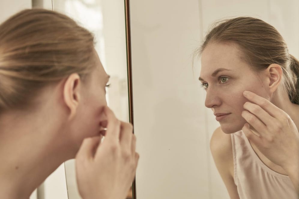 Frau betrachtet sich im Spiegel: Wenn einem Hautunreinheiten auffallen, darf man nicht mit bloßen Händen daran herumdrücken.