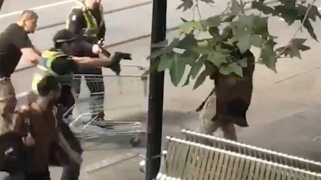 Das Standbild aus einem Video zeigt Michael Rogers (l, mit Einkaufswagen) und Polizisten, die einen Mann (r), der mit einem Messer bewaffnet ist, zu überwältigen versuchen. Das Standbild aus einem Video zeigt Michael Rogers (l, mit Einkaufswagen) und Polizisten, die einen Mann (r), der mit einem Messer bewaffnet ist, zu überwältigen versuchen.