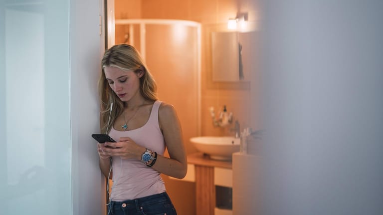 Eine junge Frau steht in der Badezimmer-Tür und schaut auf ihr Smartphone: Viele Smart Home-Geräte sind laut BSI nicht ausreichend gegen Schadsoftware geschützt.