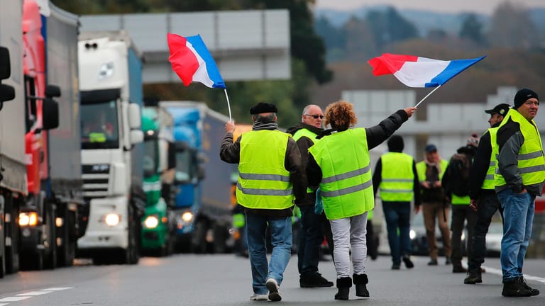 "Gelbwesten" schwenken französische Flaggen auf der Autobahn: Die französische Regierung macht den Demonstranten nun Zugeständnisse.