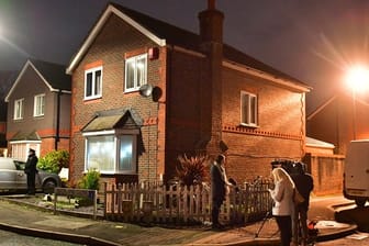 Ein Polizist und ein Kamerateam stehen vor einem Haus, das im Zusammenhang mit dem Drohnenzwischenfall am Flughafen Gatwick durchsucht wurde.