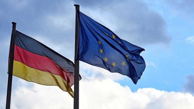 Eine Deutschland- und eine EU-Fahne vor wolkigem Himmel.