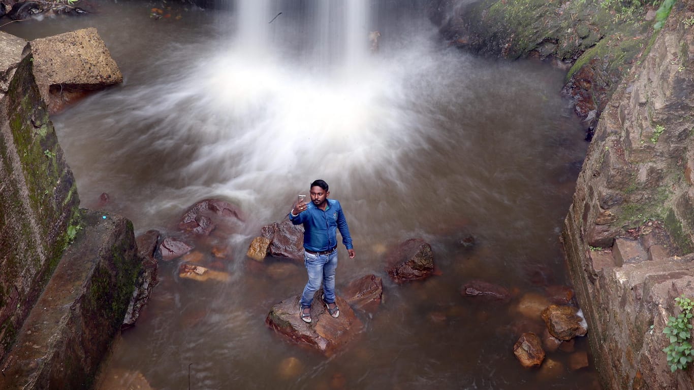 Ein indischer Tourist macht auf einem Stein stehend ein Selfie an einem Wasserfall.