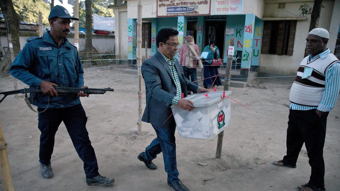 Parlamentswahl in Bangladesch: Ein Wahlhelfer trägt nach Ende der Stimmenabgabe eine Wahlurne aus einem Wahllokal. Ein bewaffneter Polizist begleitet ihn.
