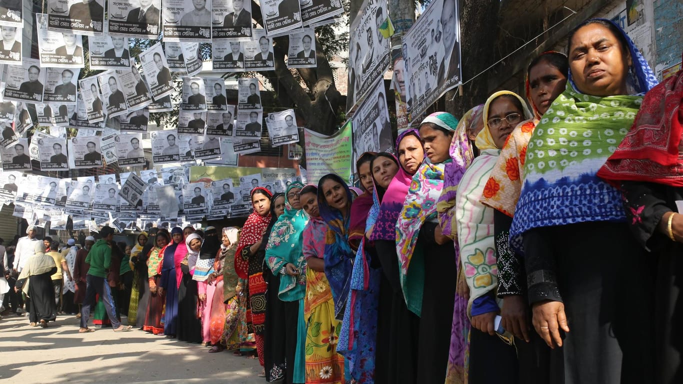 Dhaka, Bangladesch: Frauen stehen vor einem Wahllokal Schlange. Über ihnen hängen Zettel mit Wahlwerbung der Kandidaten.