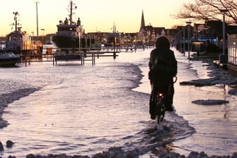 Ein Radfahrer fährt mit seinem Fahrrad die durch Hochwasser überflutete Strandpromenade von Lübeck-Travemünde entlang.