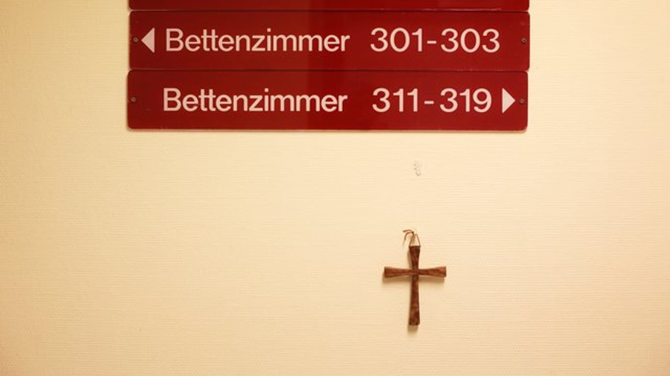 Ein kleines Holzkreuz hängt unter Hinweisschildern auf einer Station in einem kirchlichen Krankenhaus.