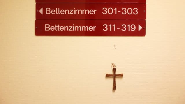 Ein kleines Holzkreuz hängt unter Hinweisschildern auf einer Station in einem kirchlichen Krankenhaus. Ein kleines Holzkreuz hängt unter Hinweisschildern auf einer Station in einem kirchlichen Krankenhaus.