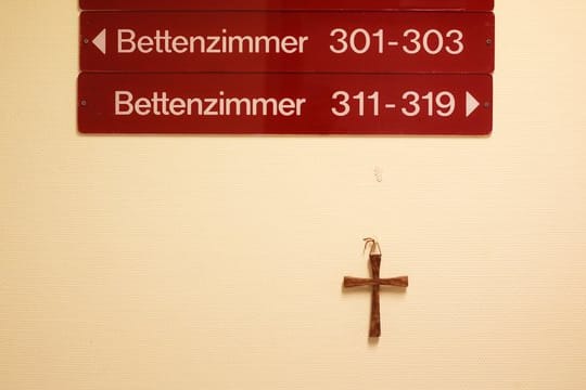 Ein kleines Holzkreuz hängt unter Hinweisschildern auf einer Station in einem kirchlichen Krankenhaus.
