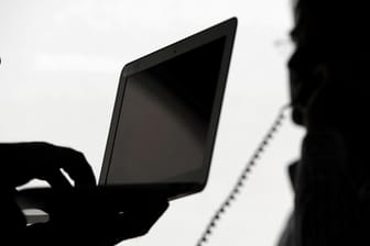 Zwei Männer stehen mit Computer und Telefon vor einem Fenster: Unbekannte Anrufer können Abzocker sein.