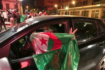 Eine Frau hält in Algier eine algerische Nationalflagge aus ihrem Auto, während im Hintergrund Menschen in den Straßen feiern.