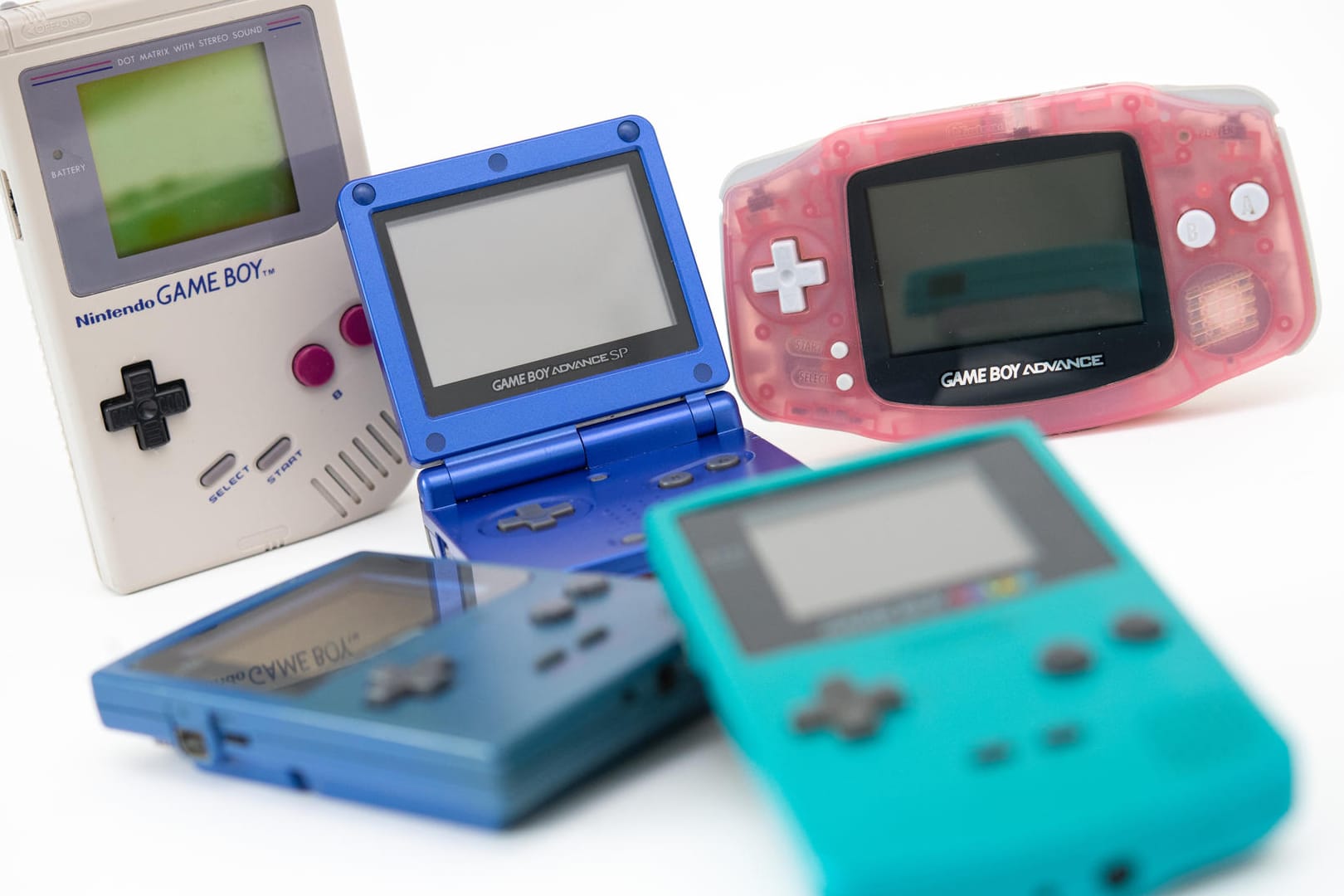 Verschieden Game-Boy-Versionen: Der erste Game Boy (l in Grau) erhielt über die Jahre etliche Nachfolger wie den Game Boy Pocket (blau, liegend), den Game Boy Advance SP (Mitte, aufgeklappt), den Game Boy Advance (r, rosa) und den Game Boy Color (vorne, mit Farbdisplay).