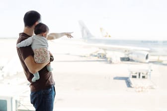 Mann mit Baby am Flughafen: In den ersten Lebensmonaten sollte abgewogen werden, ob eine Flugreise notwendig ist.