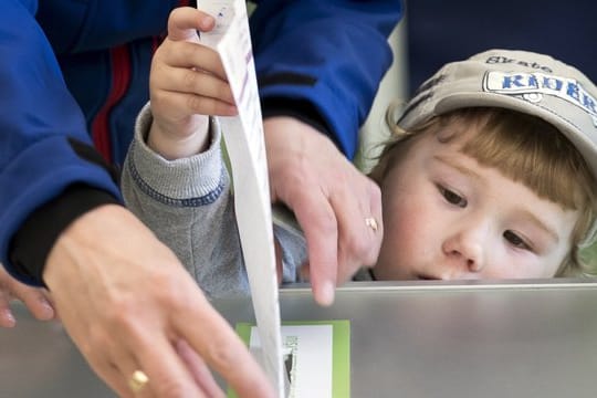 Eine Frau lässt ein Kind ihren Stimmzettel in die Wahlurne stecken.