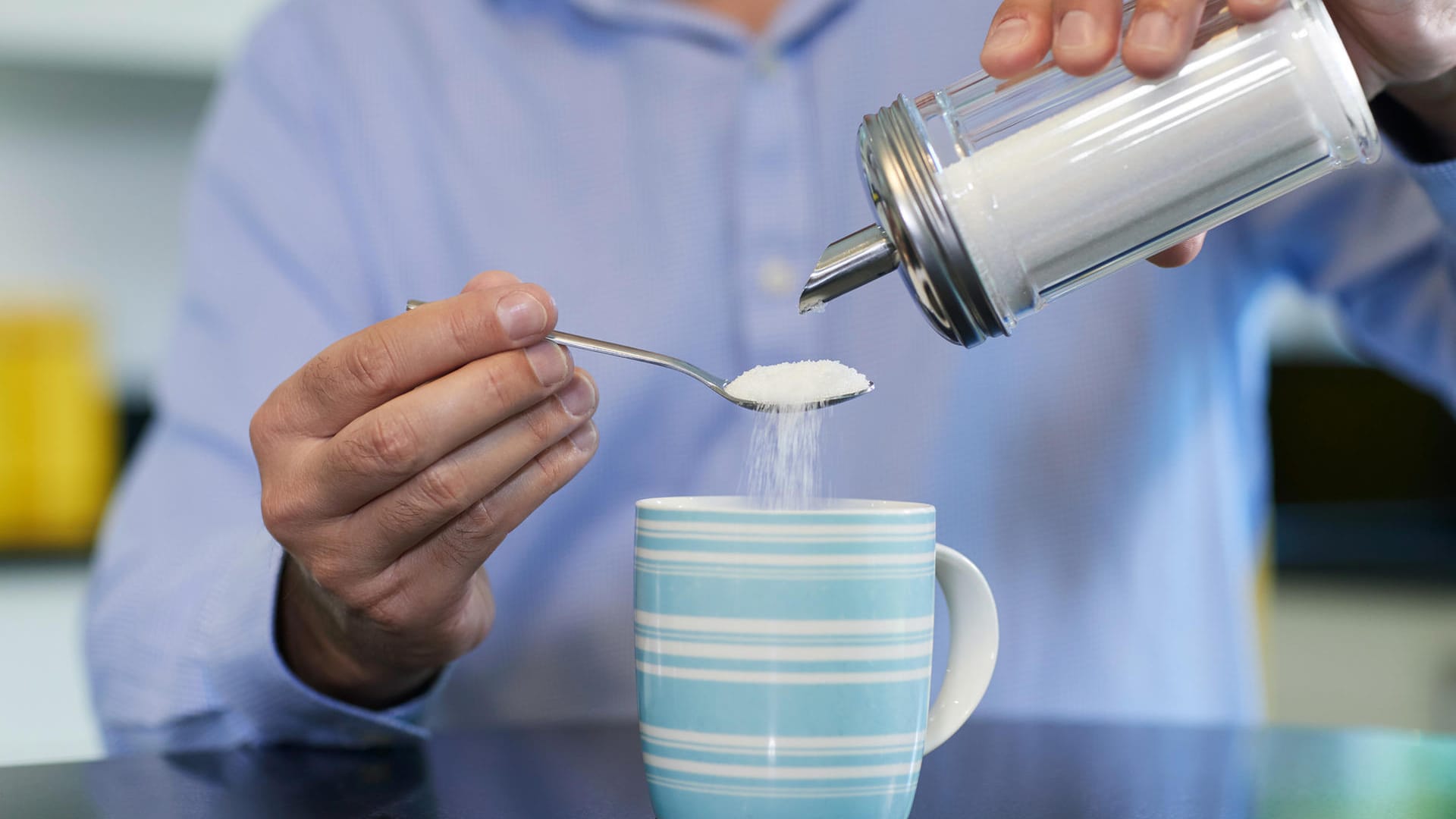 Ein Mann schüttet Zucker in seinen Kaffee: Wer seinen Zuckerkonsum verringern möchte, kann damit beginnen, statt zwei Löffel Zucker nur einen in die Tasse mit Kaffee zu rühren.