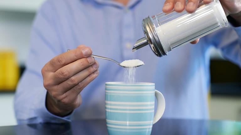 Ein Mann schüttet Zucker in seinen Kaffee: Wer seinen Zuckerkonsum verringern möchte, kann damit beginnen, statt zwei Löffel Zucker nur einen in die Tasse mit Kaffee zu rühren.