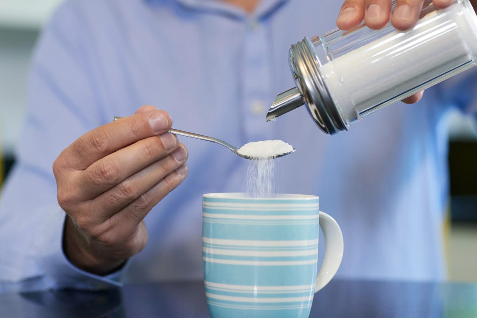 Ein Mann schüttet Zucker in seinen Kaffee: Wer seinen Zuckerkonsum verringern möchte, kann damit beginnen, statt zwei Löffel Zucker nur einen in die Tasse mit Kaffee zu rühren.