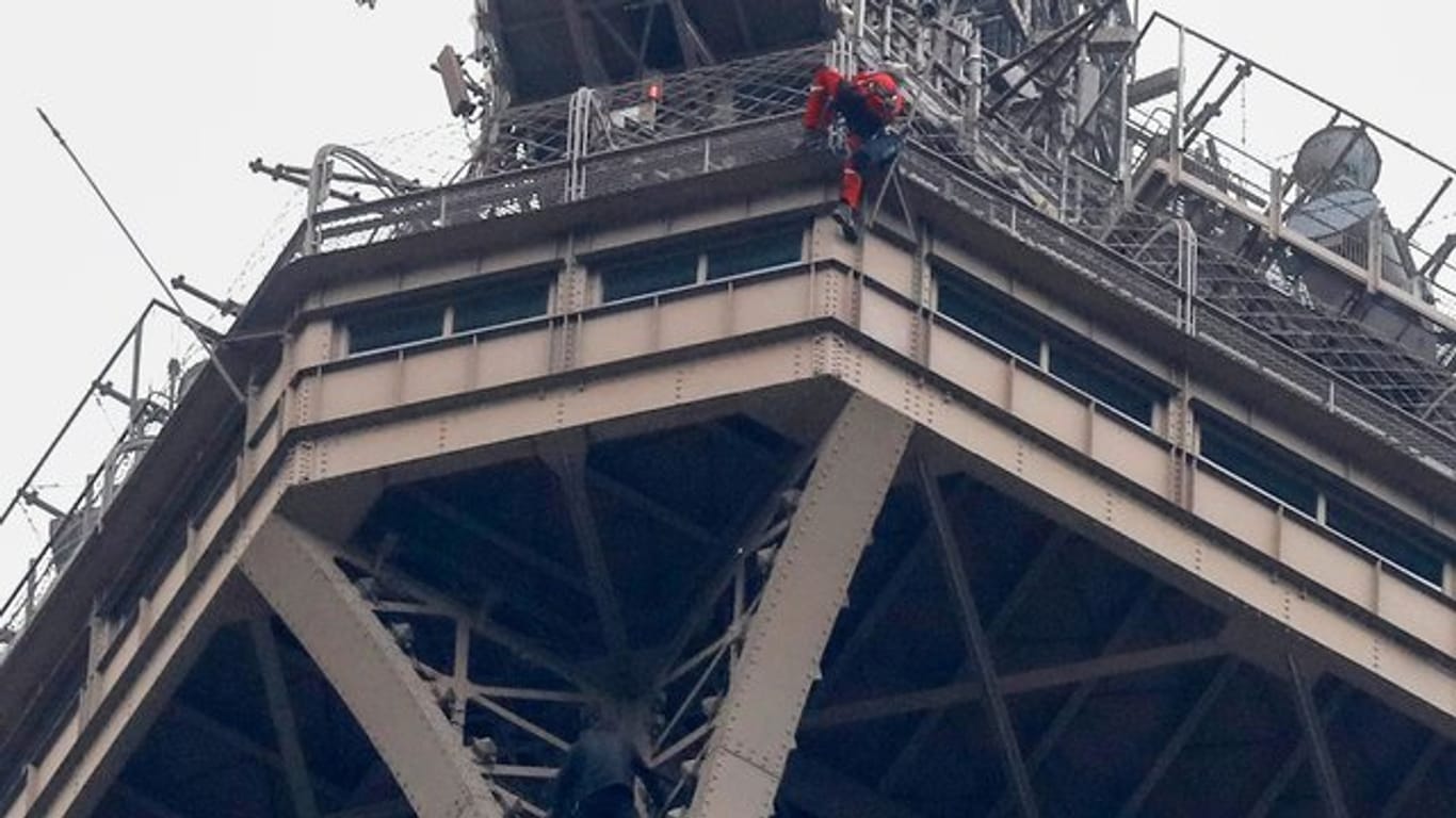 Ein Rettungshelfer (oben in roter Berufskleidung) klettert angeseilt am Eiffelturm hinunter, während unter ihm der Kletterer an die Gitterstäbe klammert.