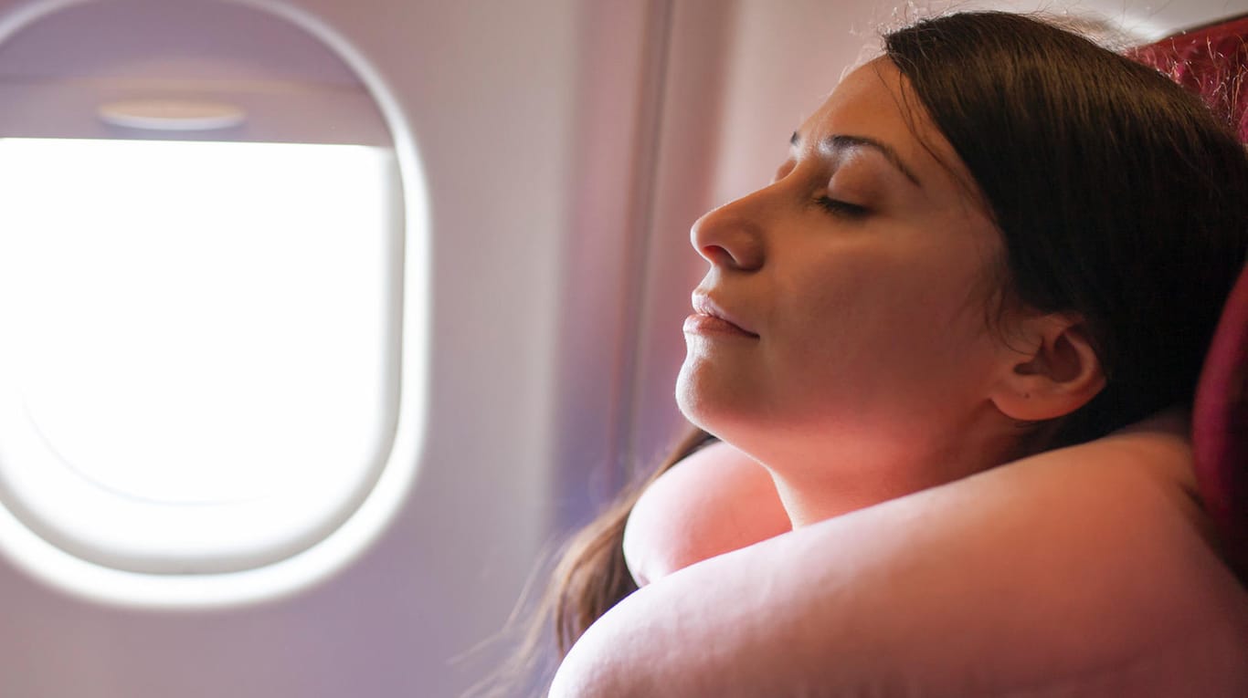 Frau sitzt mit Nackenkissen im Flugzeug: Schlafmasken und Ohrstöpsel helfen beim Einschlafen.