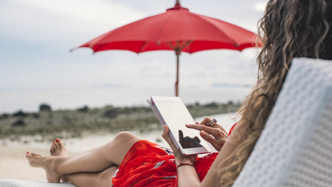 Eine Frau nutzt ihr Tablet am Strand: Mit technischen Geräten müssen Sie im Urlaub vorsichtig sein.