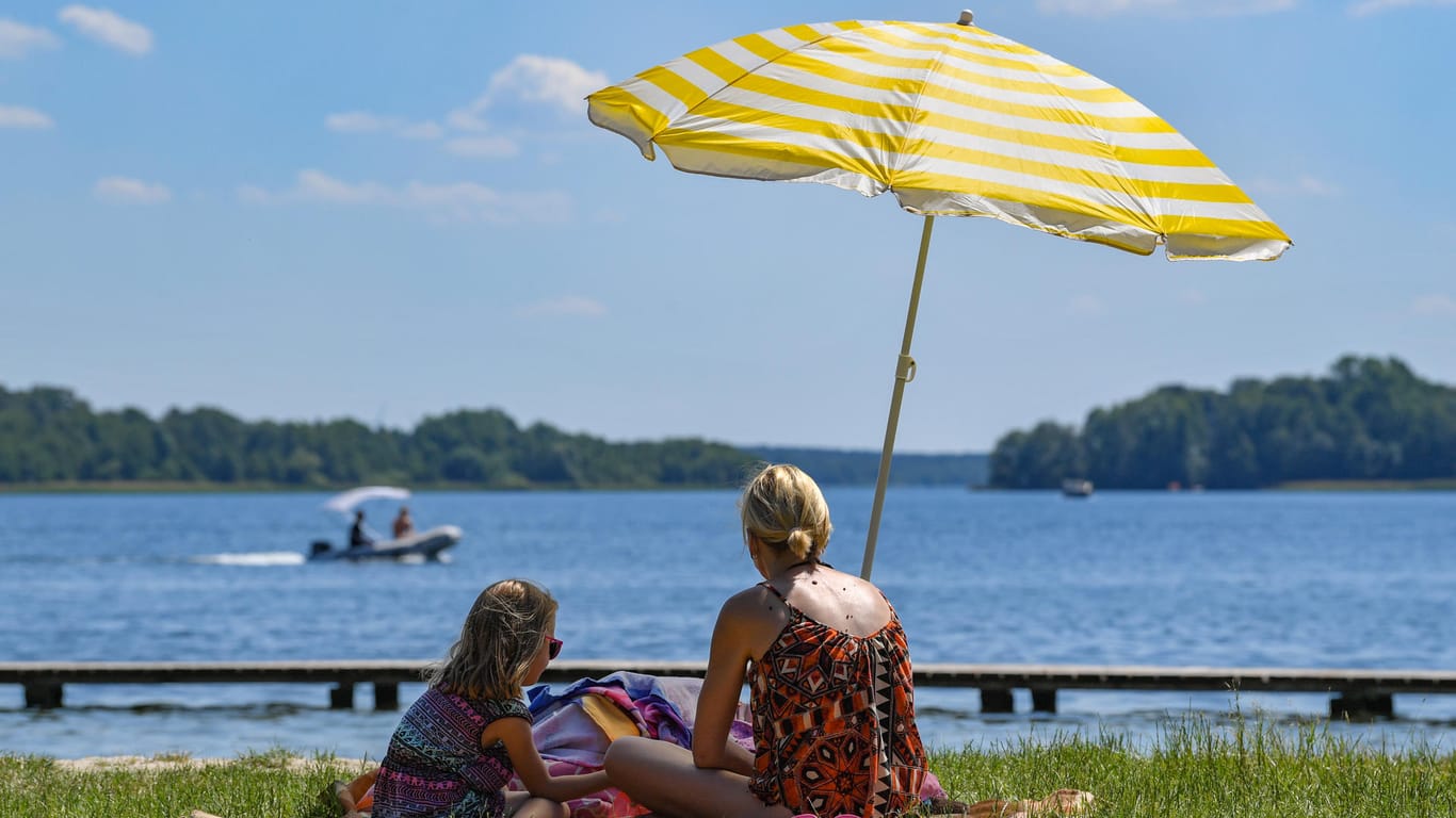 Eine Mutter und ihre Tochter sitzen unter einem Sonnenschirm im Strandbad am Storkower See: Wetterexperten rechnen damit, dass Dürreperioden zukünftig länger anhalten werden.